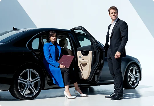 Two professionals step out of a sleek black luxury sedan in a brightly lit, minimal setting. The woman holds a portfolio while the man stands poised, creating a modern business travel scene.