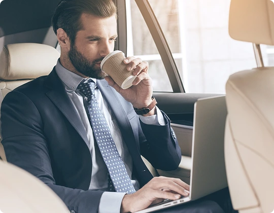 Businessman in a navy suit sipping coffee while working on a laptop in the backseat of a luxury car, with sunlight filtering through the window.