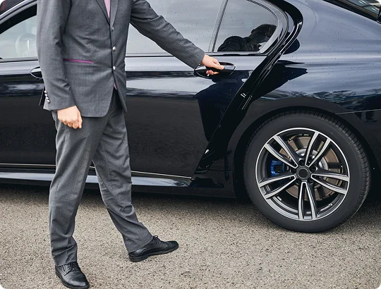 Man in a grey suit opening the rear door of a black luxury car, viewed from the side at ground level.