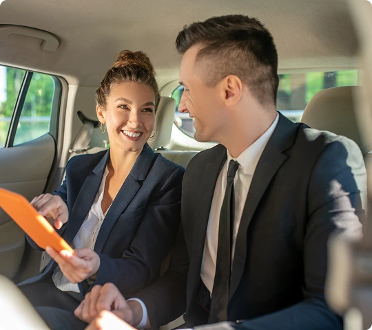 Two professionals in formal attire share a lighthearted conversation while reviewing a folder in the backseat of a premium car, capturing a moment of productive and comfortable travel.