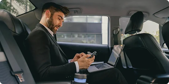 A man in a business suit is working on his phone and laptop from the backseat of a luxury car, showcasing productive travel in a sleek black interior.