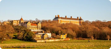 A sunlit countryside scene in Virginia featuring a stately hilltop building with red chimneys, surrounded by quaint homes, open grassy fields, and leafless forest in the background.