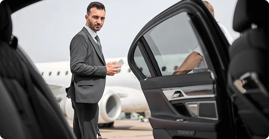 A man in a suit standing on the airport tarmac beside a private jet, holding a phone and looking toward a luxury car with its rear door open.