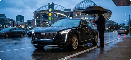 A chauffeur in a dark suit stands under a black umbrella next to a sleek, dark Cadillac sedan on a wet city street. The Lumen Field stadium is visible in the background under a blue, overcast sky.