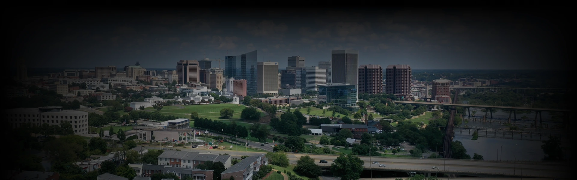 A sweeping aerial view of downtown Richmond, Virginia, shows a mix of historic buildings and modern high-rises under a moody sky, with green spaces and roadways in the foreground, illustrating the urban route from Washington DC to Virginia.