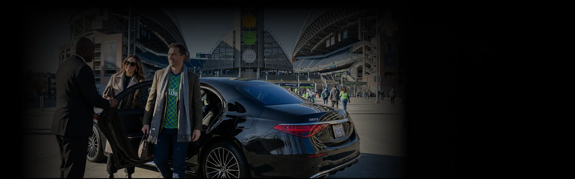 A chauffeur in a black suit holds open the door of a black luxury sedan for a smiling couple outside Lumen Field stadium.